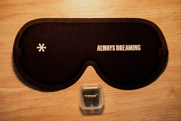 A black sleep mask with "ALWAYS DREAMING" text and a small logo, placed on a wooden surface.