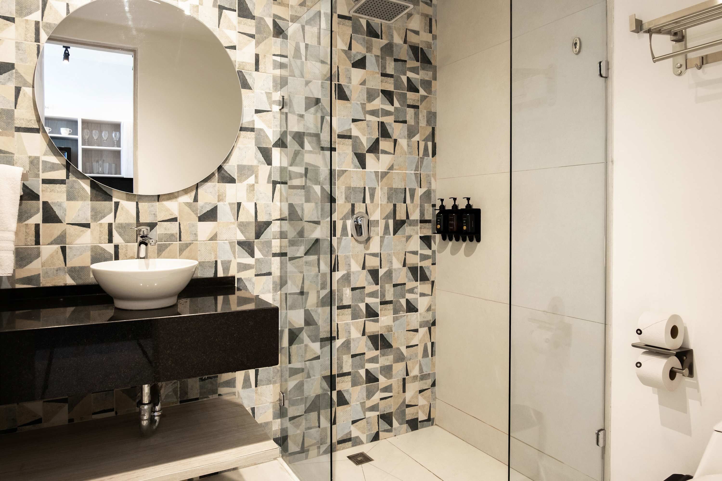 Un bagno moderno con pannelli decorativi in piastrelle geometriche, uno specchio rotondo, un lavandino bianco e una cabina doccia in vetro.