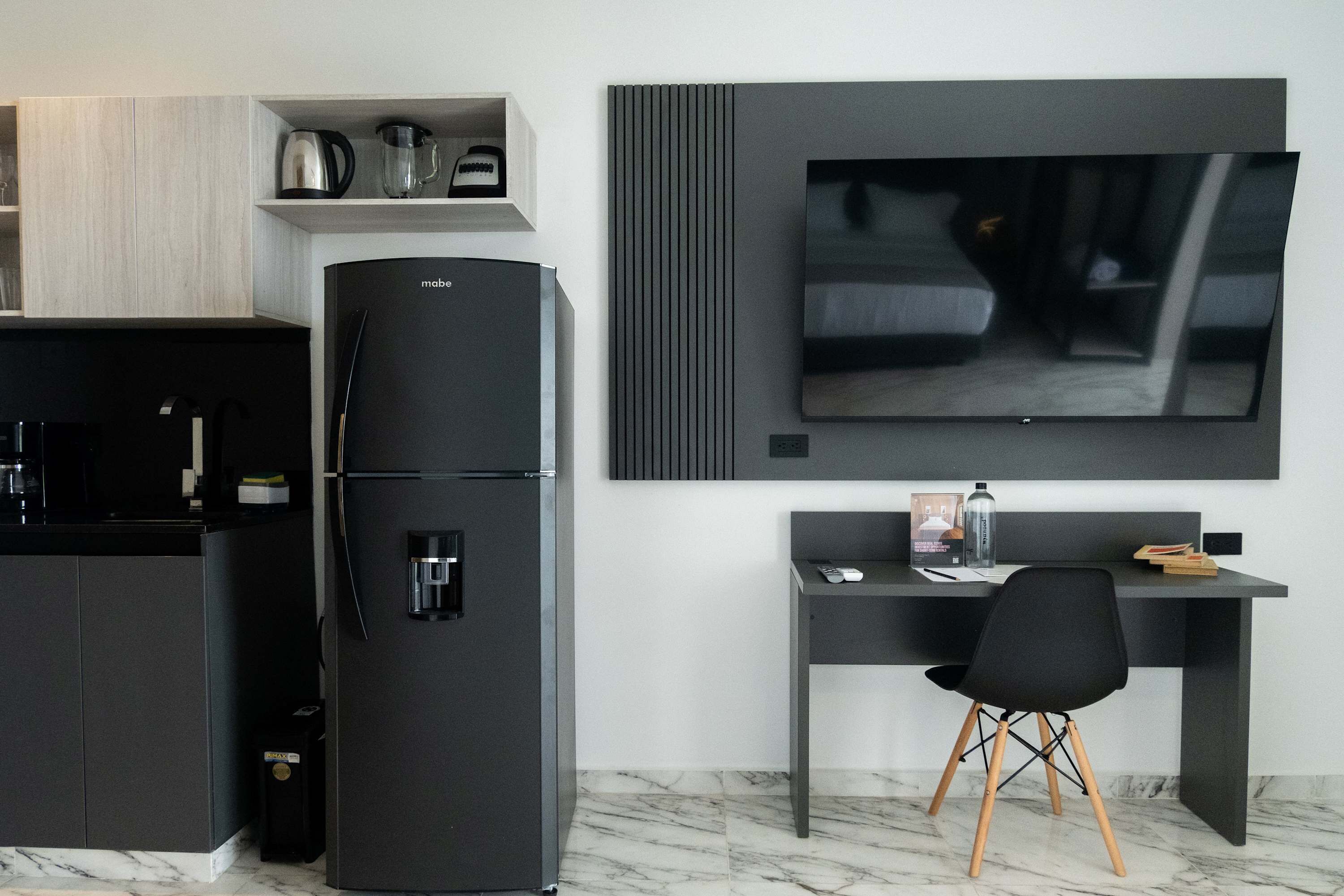 A modern kitchen with a black refrigerator, a coffee maker, and a kettle on the countertop. There is a black chair and a desk with a flat-screen TV mounted on the wall.