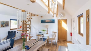 Um quarto amplo com escada de madeira, área de jantar com mesa e cadeiras de madeira, pequena escrivaninha com luminária e uma grande janela que proporciona iluminação natural.
