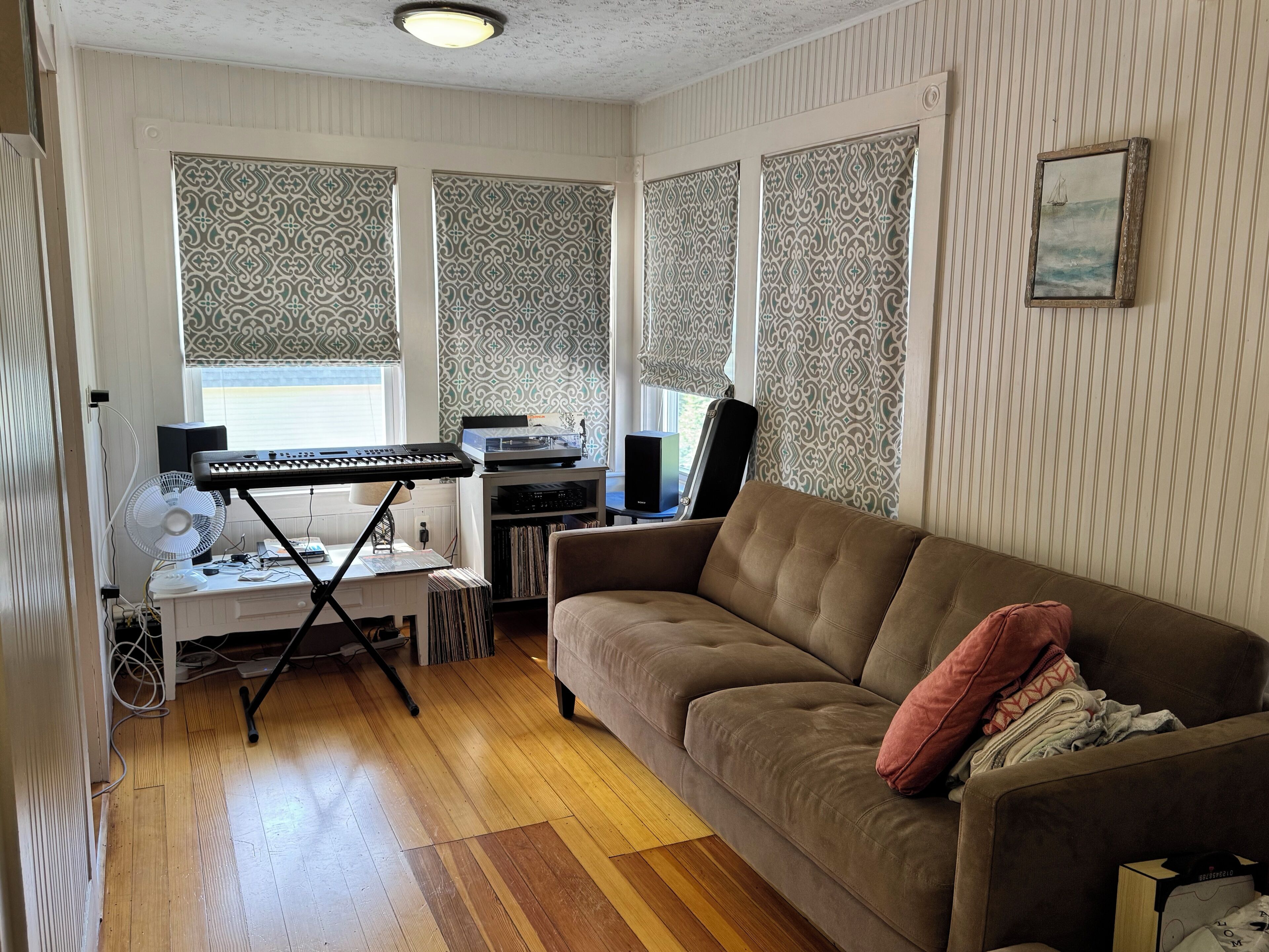 A living room with a brown sofa, a small table with a keyboard and speakers, a fan, and a bookshelf with vinyl records.