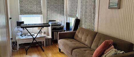 A living room with a brown sofa, a small table with a keyboard and speakers, a fan, and a bookshelf with vinyl records.
