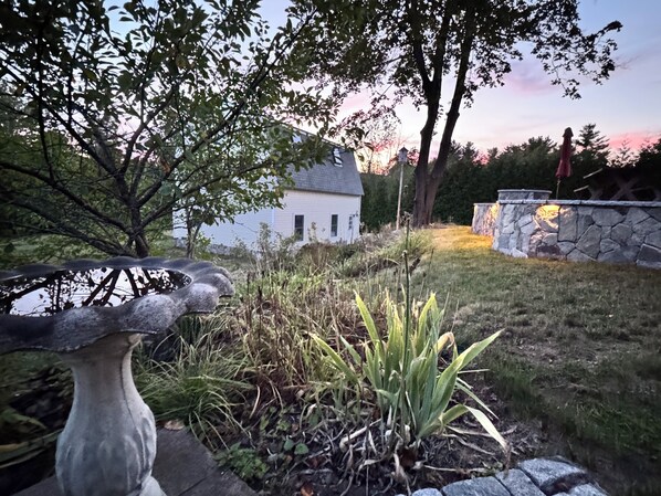A garden with a stone sculpture, a white house, and a stone wall. - Bright 3BR Coastal Home • Fire Pit, Outdoor Shower, EV Charger• Near York Harbor (York)