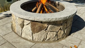 A stone fire pit with logs burning, surrounded by stone steps and a tree. - Bright 3BR House • New patio, Fire Pit, Outdoor Shower, EV Charger • Beach 4min (York)