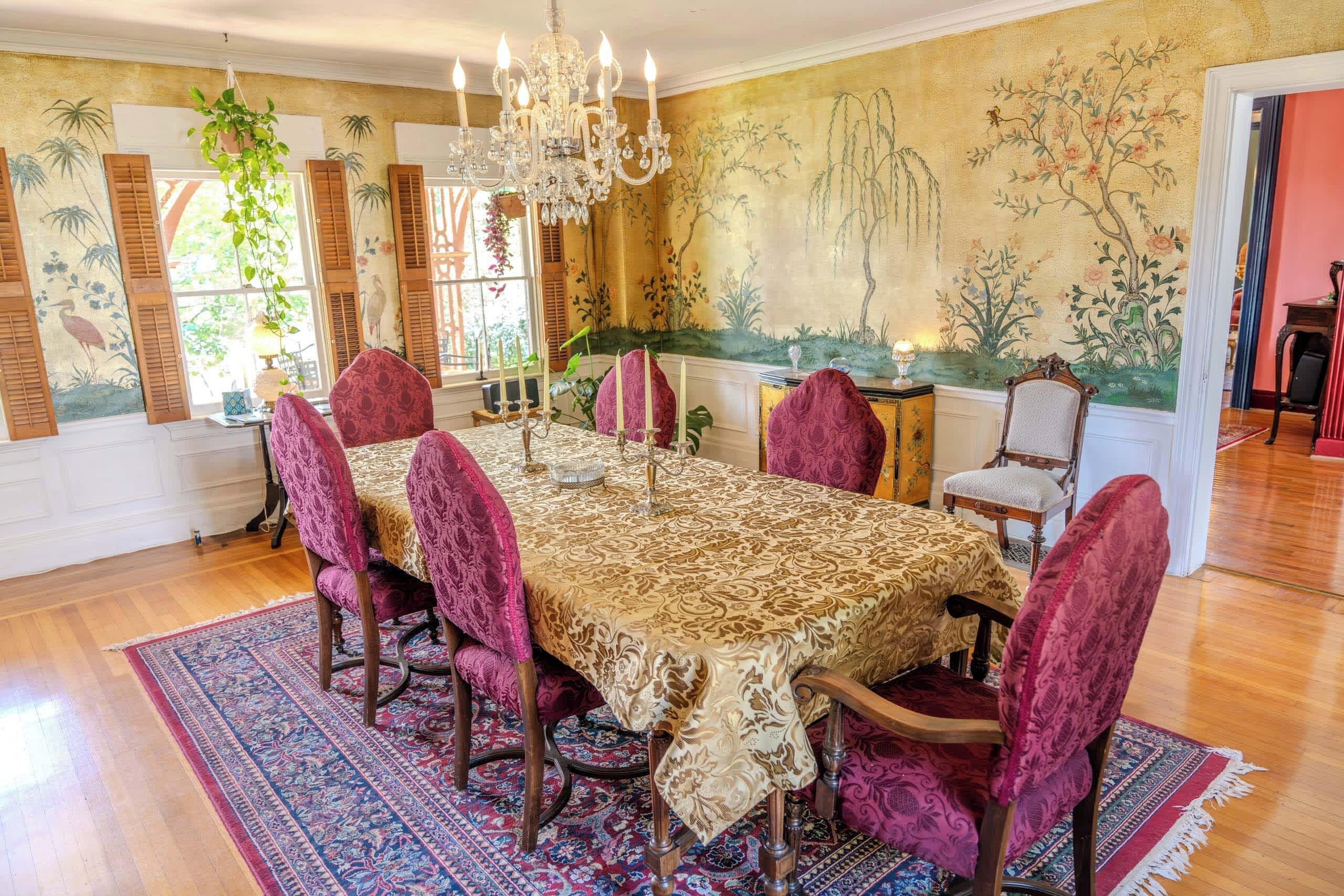 Une salle à manger avec une grande table, des chaises décorées, un lustre et une fresque murale.