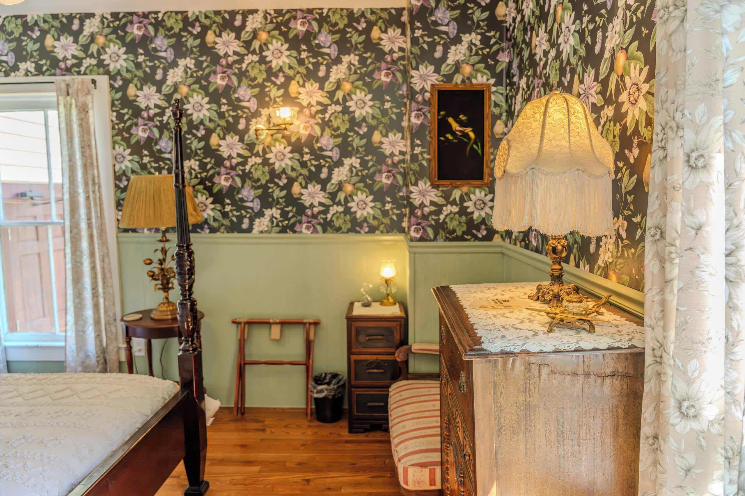 A bedroom with floral wallpaper, a wooden bed, a nightstand, a lamp, and a dresser.