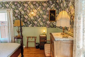 Une chambre à coucher avec un papier peint à motifs floraux, un lit en bois, une table de chevet, une lampe et une commode.