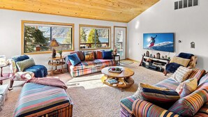 Un salon chaleureux avec un plafond en bois, de grandes fenêtres, un ensemble de canapés aux couleurs vives, une table basse et un téléviseur à écran plat.