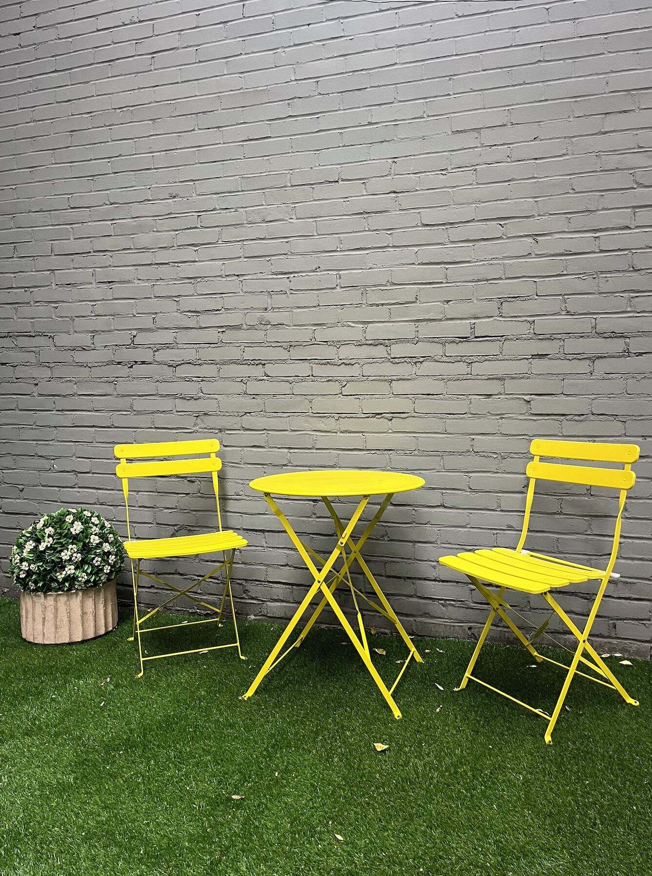 Un conjunto de sillas amarillas y una mesa frente a un muro de ladrillo, con una planta en maceta sobre el césped.