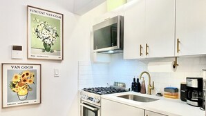 Moderni keittiö, jossa on valkoiset kaapit, ruostumattomasta teräksestä valmistetut kodinkoneet, mikroaaltouuni, liesi ja astianpesukone. Seinällä on kehystettyjä tauluja ja lattialla musta matto.
