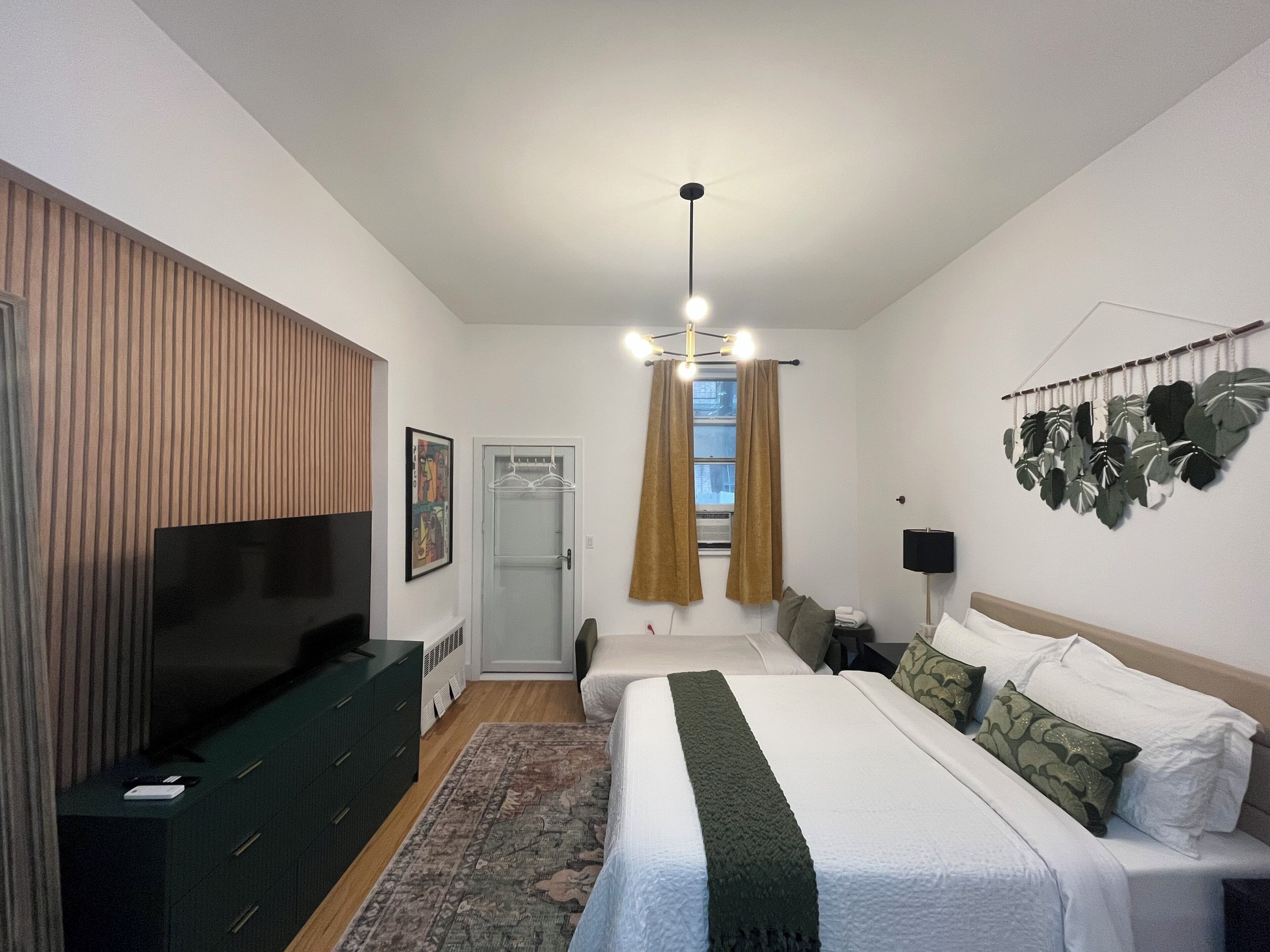 A bedroom with a bed, a TV, a dresser, a window with curtains, and a wall-mounted plant.