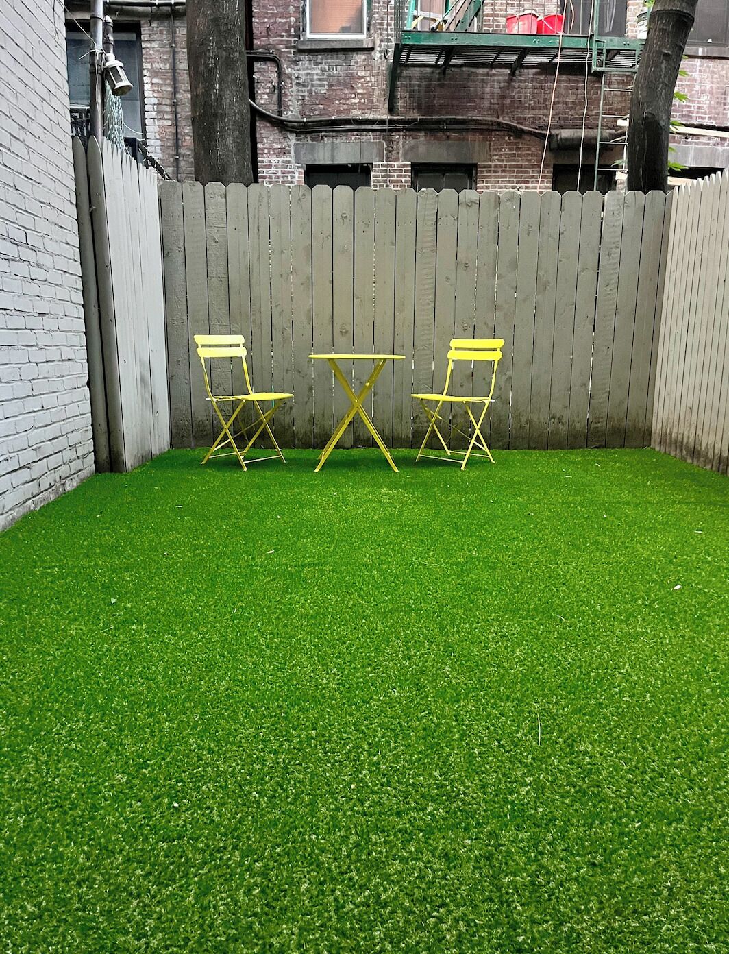 Een omheinde achtertuin met kunstgras, een kleine gele tafel en twee gele stoelen.