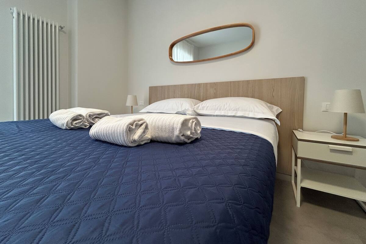 A neatly made bed with a blue quilt, two rolled towels, a wooden headboard, a bedside table with a lamp, and a mirror on the wall.
