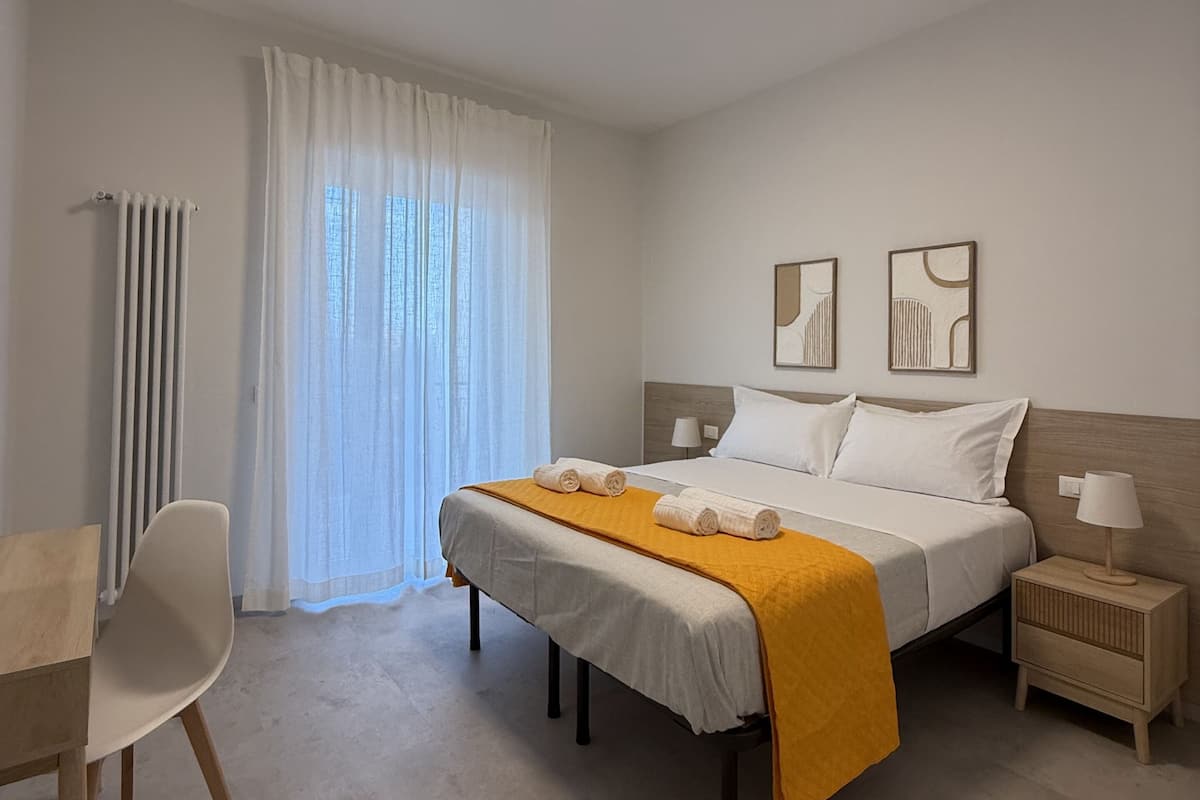 A modern hotel room with a neatly made bed, a bedside table with a lamp, a chair, and a radiator.