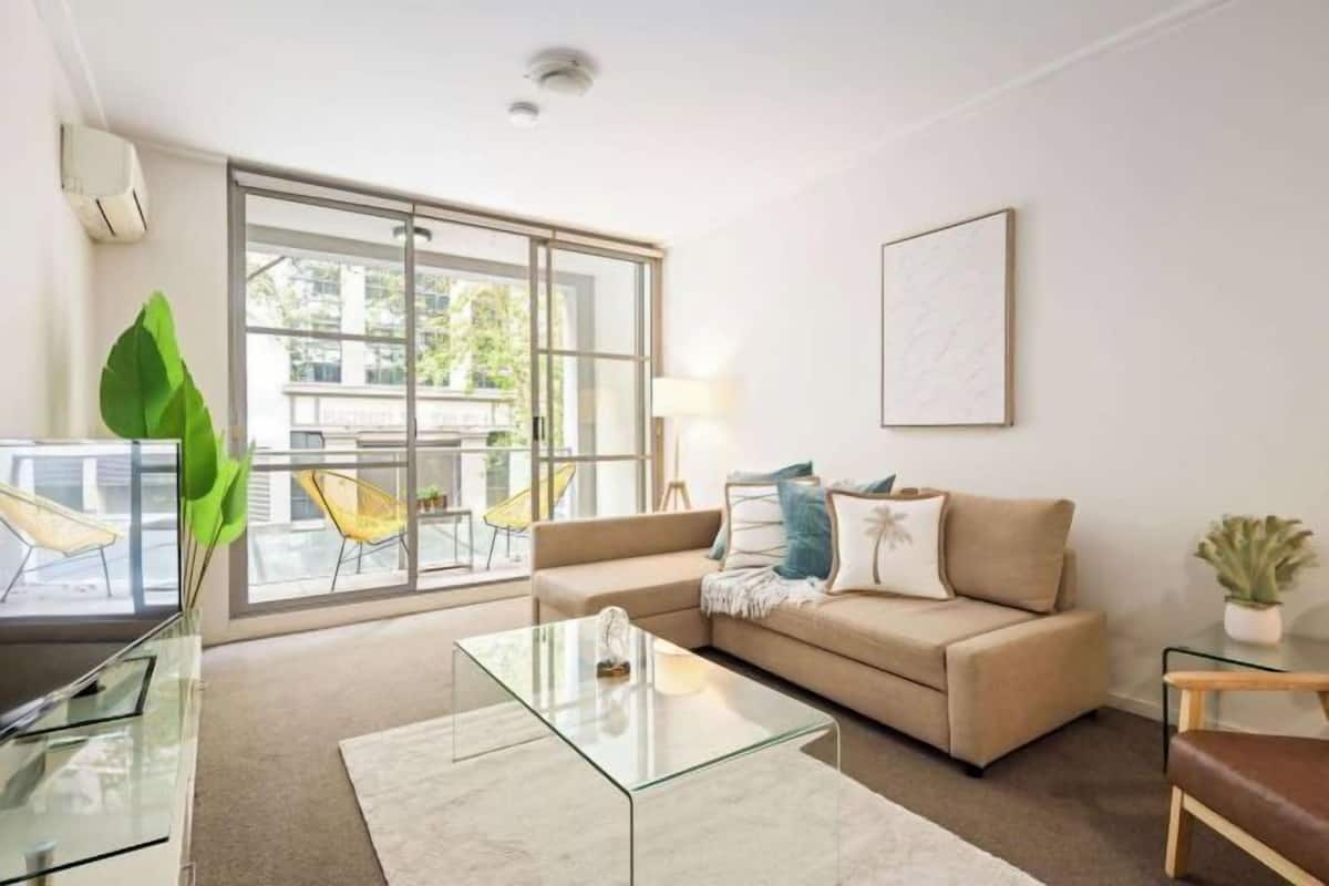 A modern living room with a glass coffee table, beige sofa, and a large window with a view of outdoor furniture.
