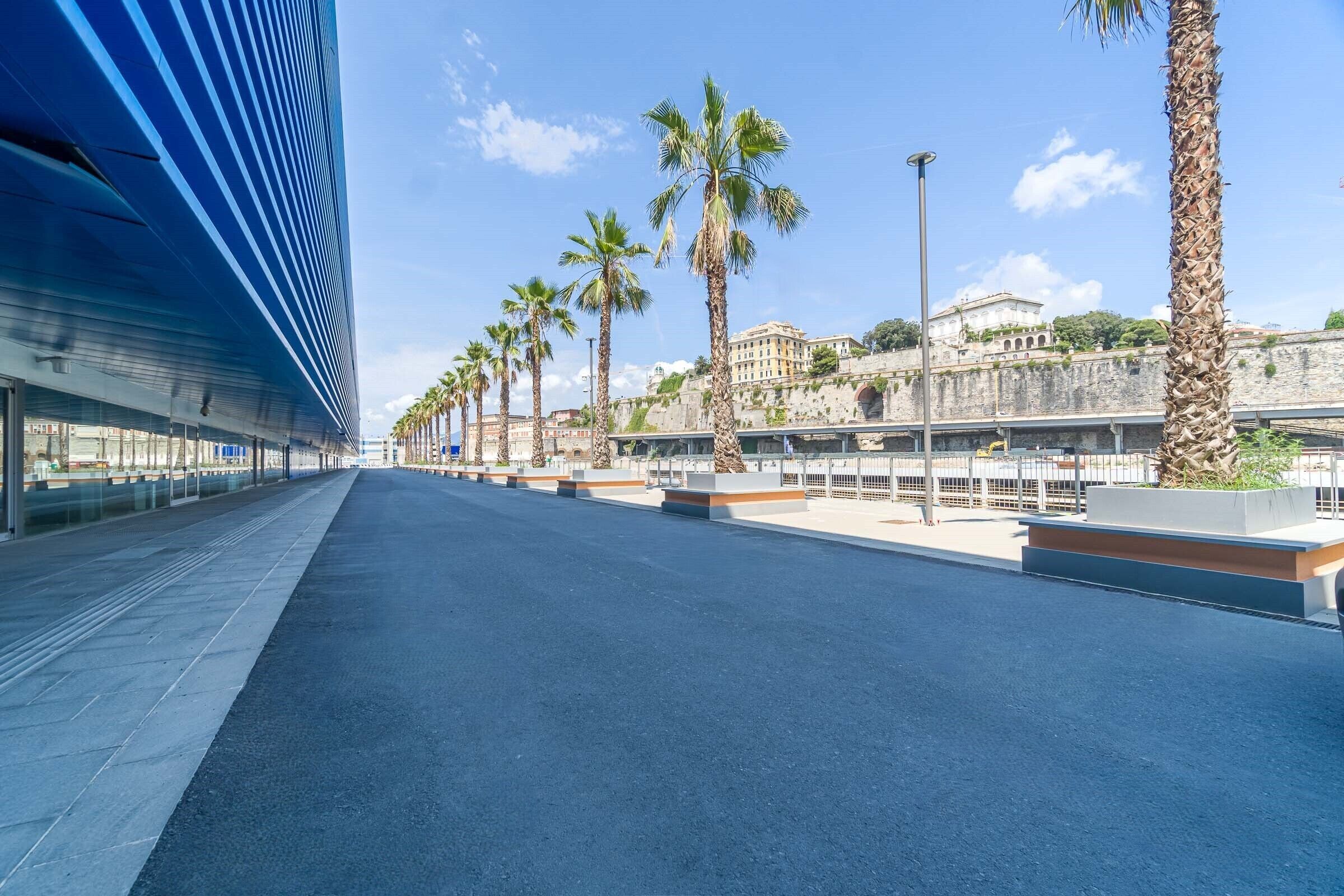 A modern building with a blue facade and a long, empty street lined with palm trees and a historic stone wall.