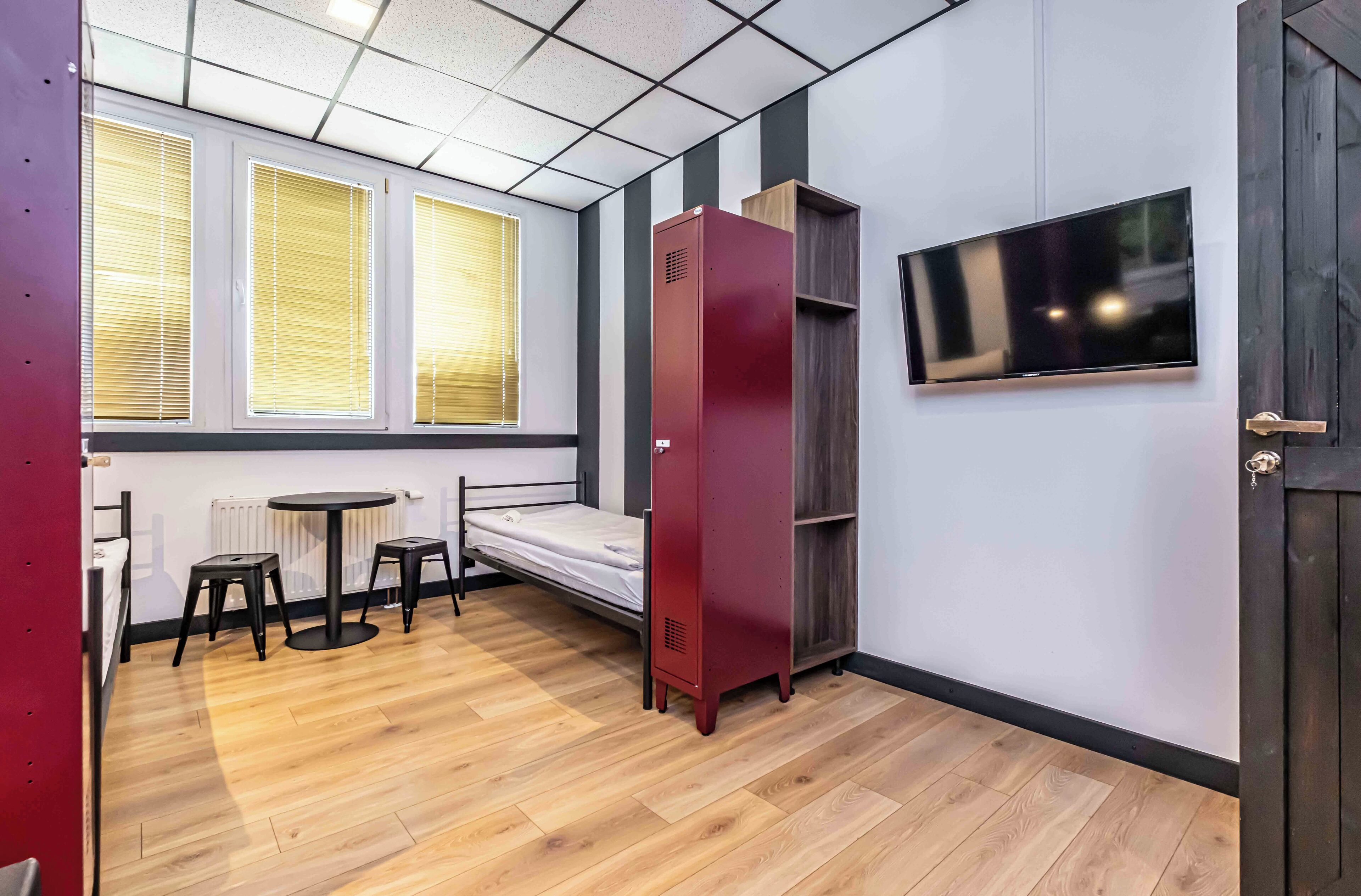 Habitación compacta con cama, una mesita, dos taburetes, un armario rojo, televisor montado en la pared y ventana con persianas.