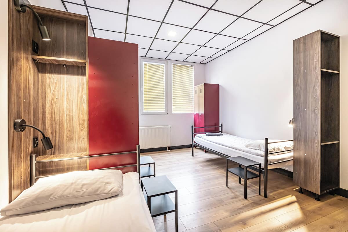 A modern dormitory room with two beds, a wooden wardrobe, a desk, and a red accent wall.