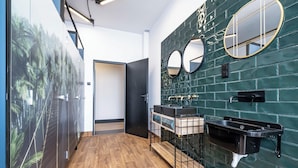 A modern bathroom with a green tiled wall, two round mirrors, a wooden floor, and a wooden bench. - VOLA Hostel (Warsaw)