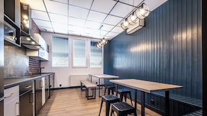 A modern kitchen with wooden countertops, a built-in stove, and a window with blinds. There are two tables and stools for dining. - VOLA Hostel (Warsaw)