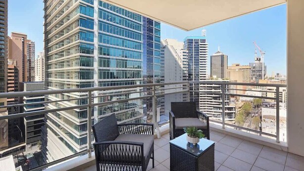 Varanda com vista para edifícios altos, guarda-corpo de vidro e móveis de exterior.