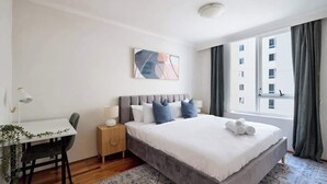 Quarto com cama, mesas de cabeceira, escrivaninha e janela com cortinas.