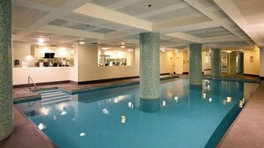 Uma grande piscina interior com água azul cristalina, rodeada por paredes e colunas revestidas em azulejos, e um teto com iluminação embutida.