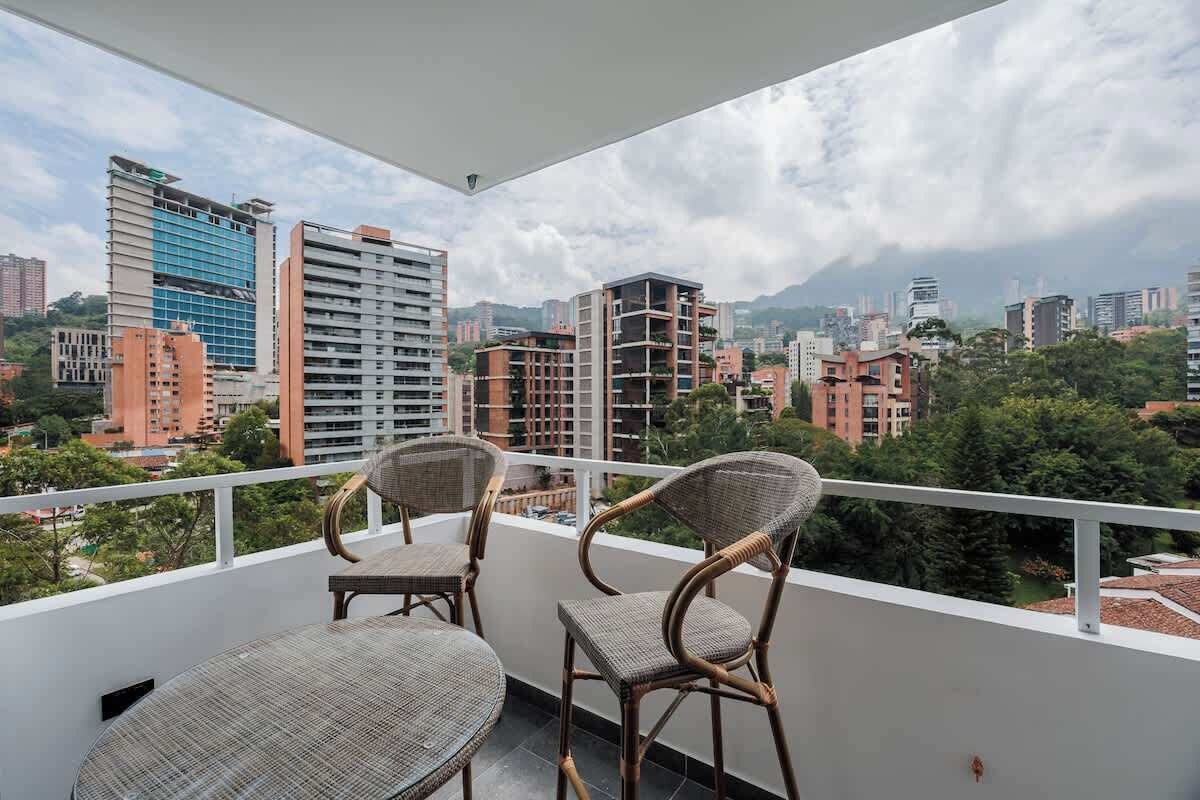 A balcony with wicker chairs and a table, overlooking a cityscape with various buildings and greenery.