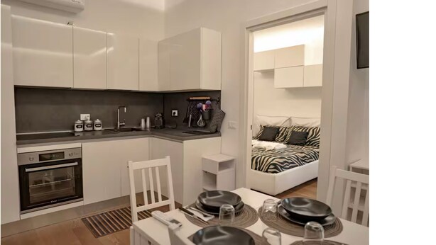 A compact kitchen with white cabinets, a built-in oven, and a dining area with a table set for two. - COZY NEW LOFT-CITYLIFE (Milano)