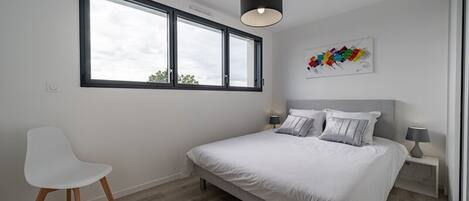 A bedroom with a bed, a chair, a window, and a wall art piece.