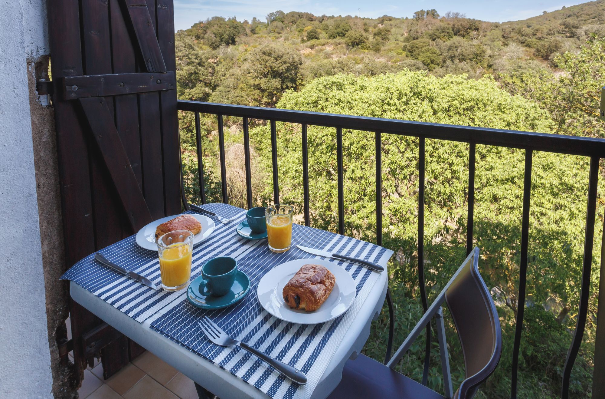 A balcony table with breakfast items, including croissants, a glass of orange juice, and a cup, overlooking a lush green landscape.