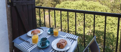A balcony table with breakfast items, including croissants, a glass of orange juice, and a cup, overlooking a lush green landscape.