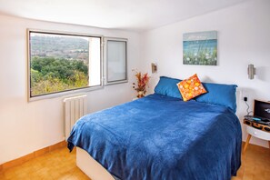 A bedroom with a large bed, a window with a view of greenery, a wall-mounted light, and a small table with a speaker.