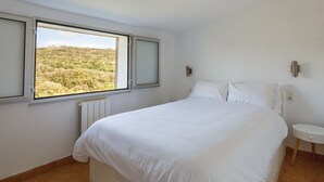 A bedroom with a large bed, a small bedside table, and a window with a view of a hilly landscape. - Cosy Apartment in Ancient Gabian with River and Vineyard Views (Gabian)