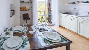 A dining area with a table set for four, a kitchen with white cabinets, and a view of the outdoors through a sliding door. - Cosy Apartment in Ancient Gabian with River and Vineyard Views (Gabian)