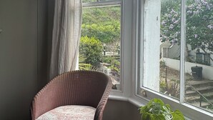 A room with a wicker chair, a small wooden table with a plant, and a view of a garden through a large window. - Peaceful Hastings old town hideaway (Hastings)