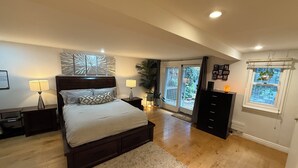 A bedroom with a large bed, two bedside tables with lamps, a wooden headboard, a large window with a view, and a sliding glass door leading to a balcony.