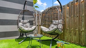 Two hanging chairs with cushions, a small round table, and a glass of drink on a grassy area with a wooden fence in the background.