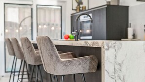 A modern kitchen with a marble countertop, a black backsplash, and a stainless steel faucet. There are three bar stools with quilted upholstery.