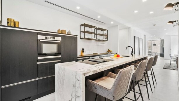 A modern kitchen with a marble island, dark cabinetry, and a dining area with bar stools.