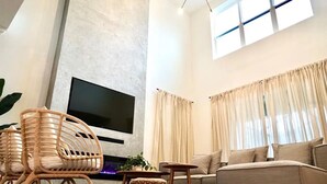 Ein modernes Wohnzimmer mit einem flachbildfernseher an einer strukturierten Wand, einem Sofa, einem Korbstuhl und einem kleinen Tisch mit einer Pflanze.