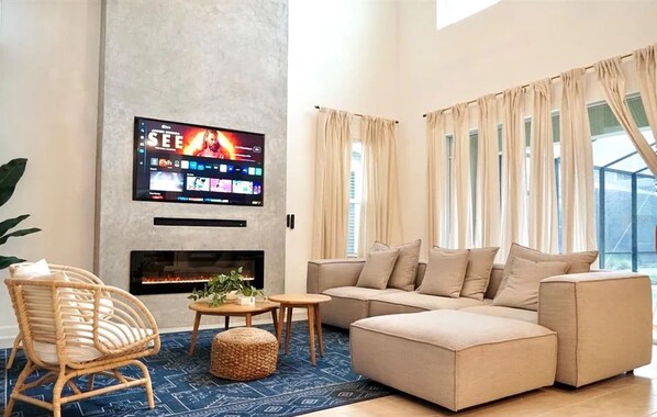 Ein modernes Wohnzimmer mit einem an der Wand montierten Flachbildfernseher, einem Kamin, einem beigefarbenen Sofa und einem großen Fenster mit Vorhängen.