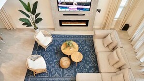 Ein Wohnzimmer mit einem beigen Sofa, zwei runden Couchtischen aus Holz, einer großen Kühlpflanze und einem Flachbildfernseher, der über einem Kamin angebracht ist.