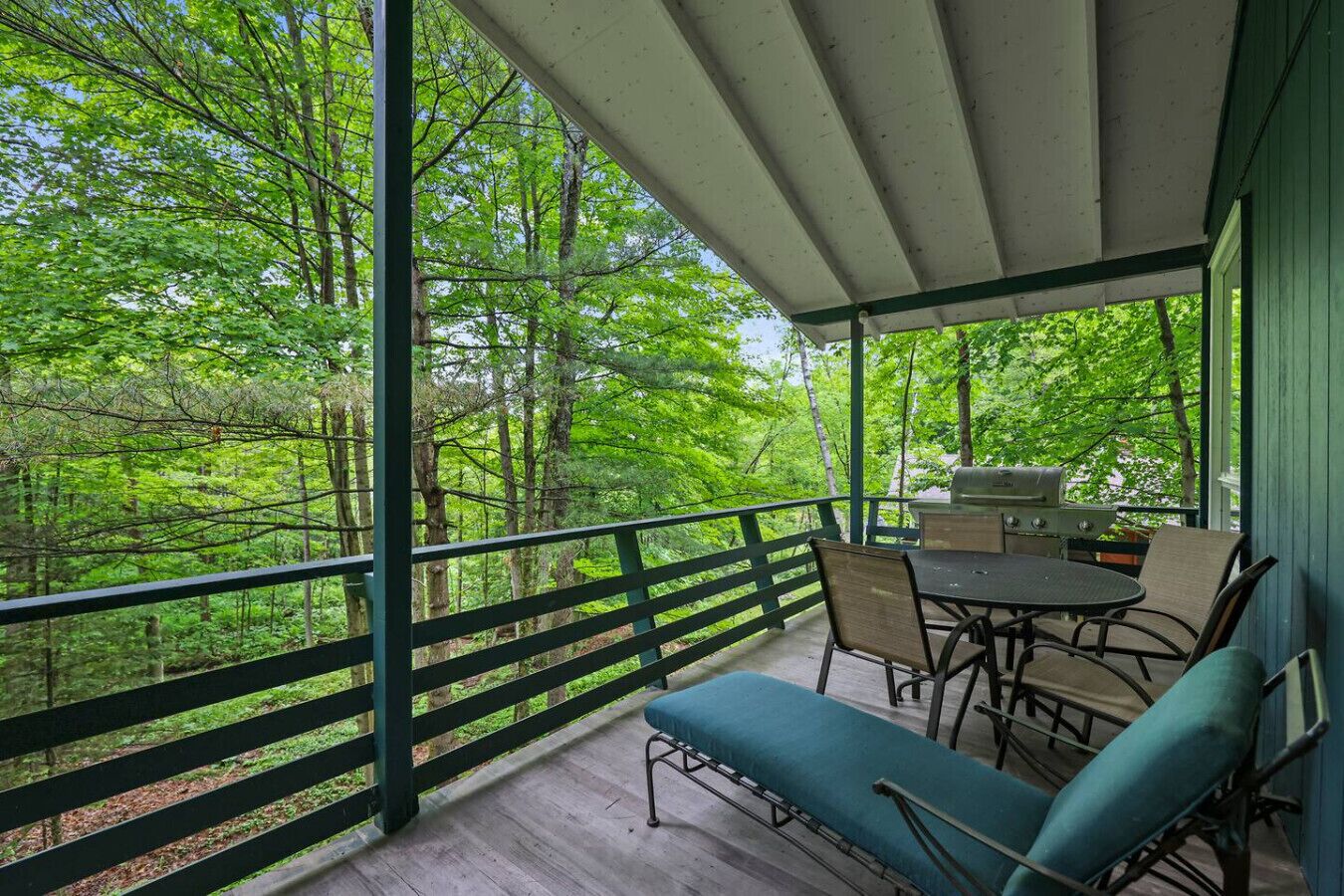 A covered outdoor area with a table and chairs, surrounded by a wooden deck and a green railing.