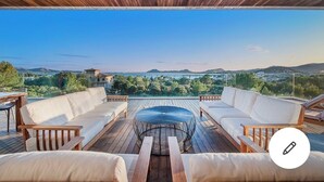 A wooden deck with a round table and sofas, overlooking a scenic landscape with hills and a bridge.