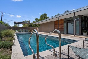 A modern outdoor pool area with a clear blue pool, metal pool ladders, lounge chairs, and a covered seating area.
