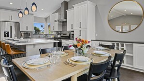 A modern kitchen with white cabinets, a central island, and a dining area with a wooden table set for four. - Pacific Beach Retreat Coastal Getaway w/ Pool (San Diego)
