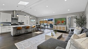 A modern kitchen with white cabinets, a central island with bar stools, and a dining area with a large window. - Pacific Beach Retreat Coastal Getaway w/ Pool (San Diego)