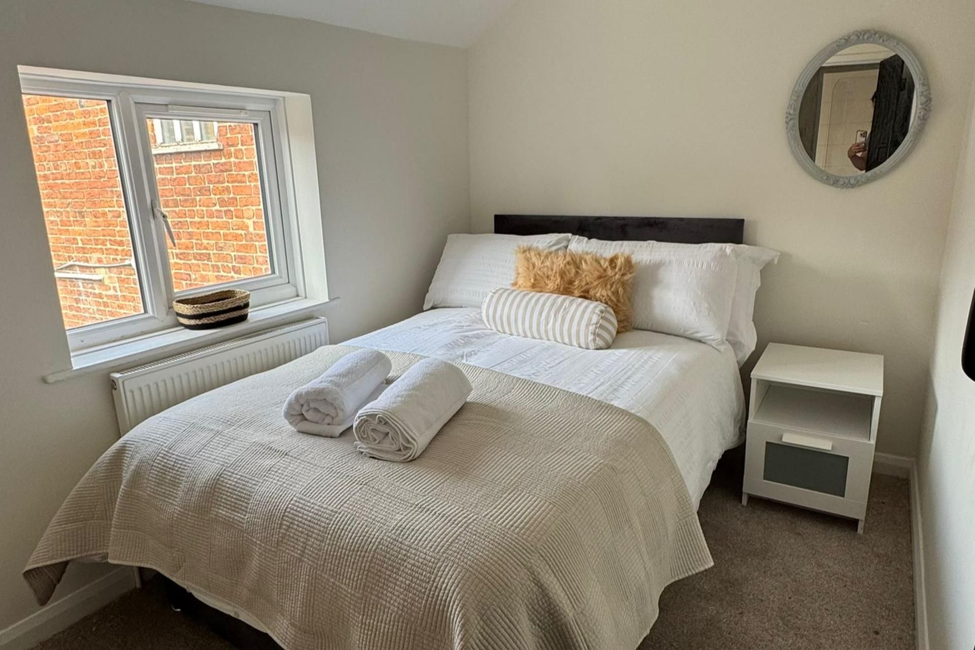 A neatly made bed with white linens and rolled towels, a bedside table with a lamp, and a window with a view of a brick wall.