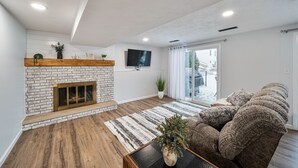 Een woonkamer met een metselwerk openhaard, een flatscreen televisie aan de muur, een bruine bank en een houten vloer.